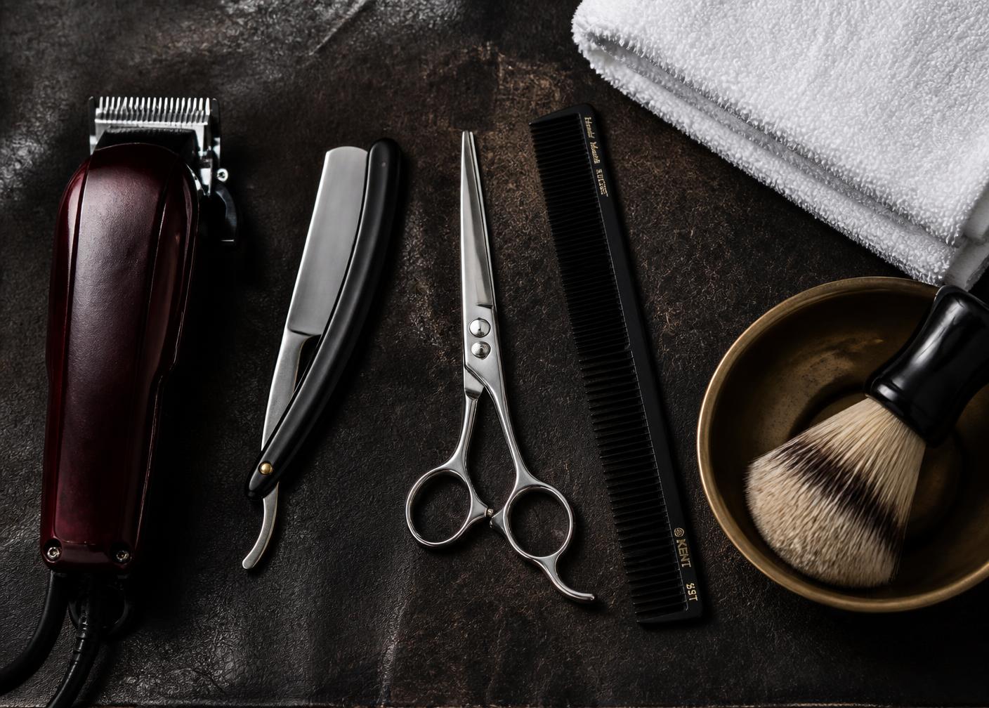 Clippers, straight razor, shears, comb, folded white towel and a brass shaving bowl laid out on dark leather